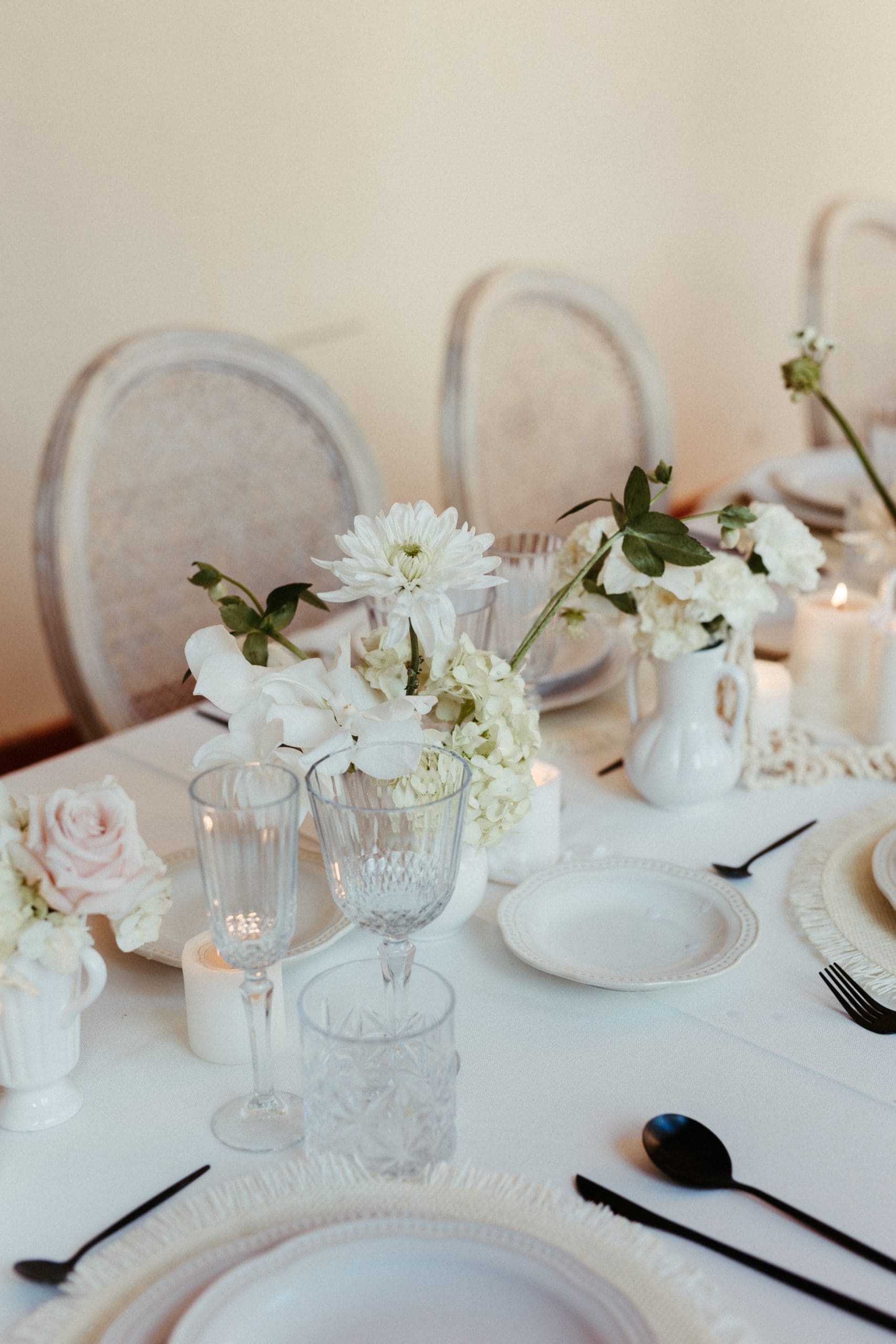 Mesa blanca decorada con pequeños jarrones con flores blancas y cubertería negra en la Cartuja de Ara Christi, Valencia. Creada por wedding planner de Bouclé Weddings.