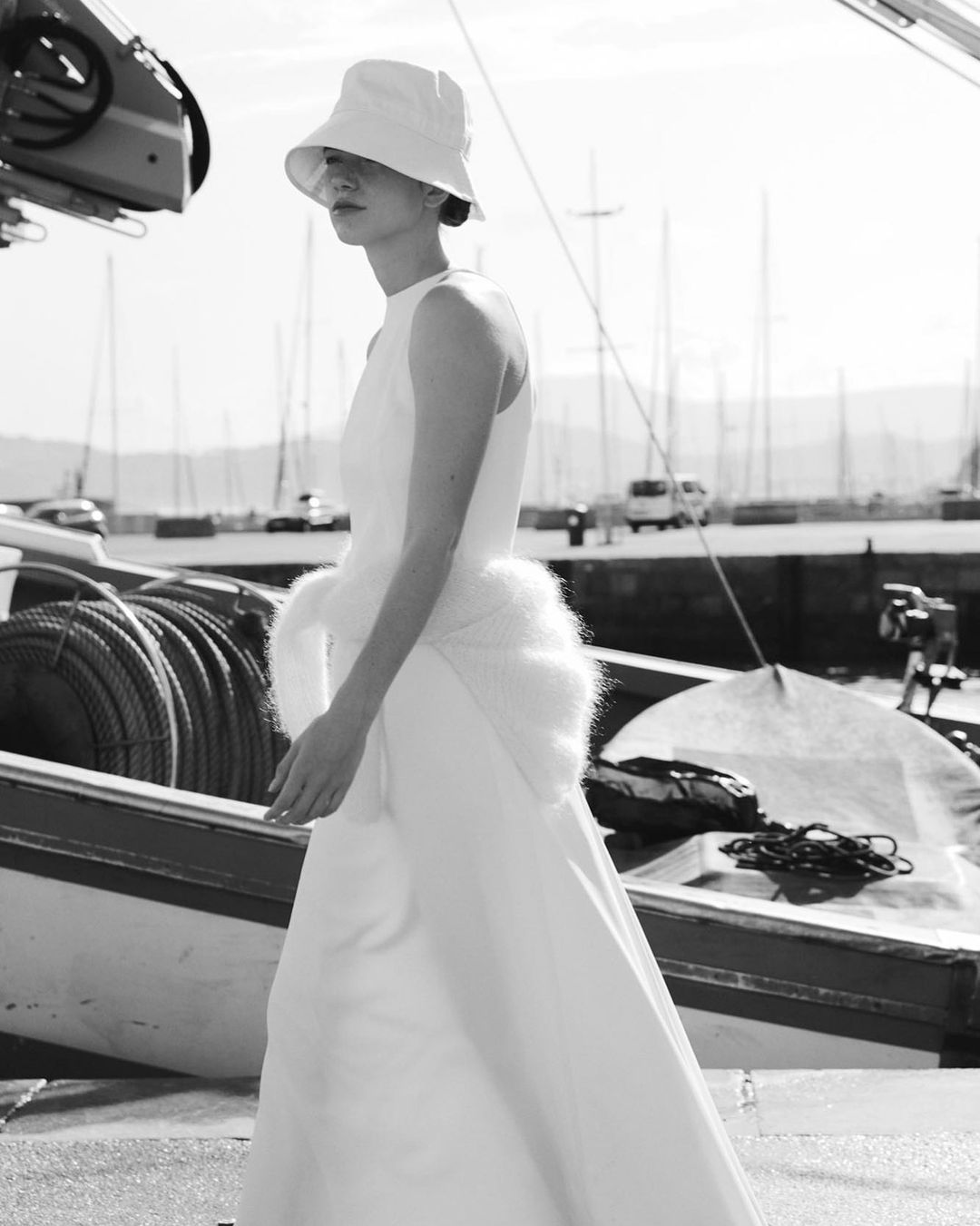Novia con gorro blanco vistiendo un vestido de Claudia Llagostera caminando por un puerto marítimo.