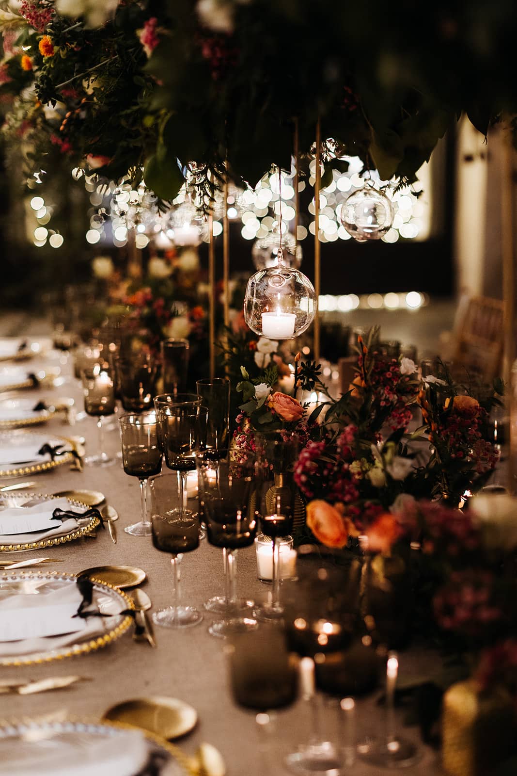 Mesa de boda con vajilla dorada, lámparas colgantes y abundantes flores en tonos verdes y rojos, creando una atmósfera romántica y lujosa
