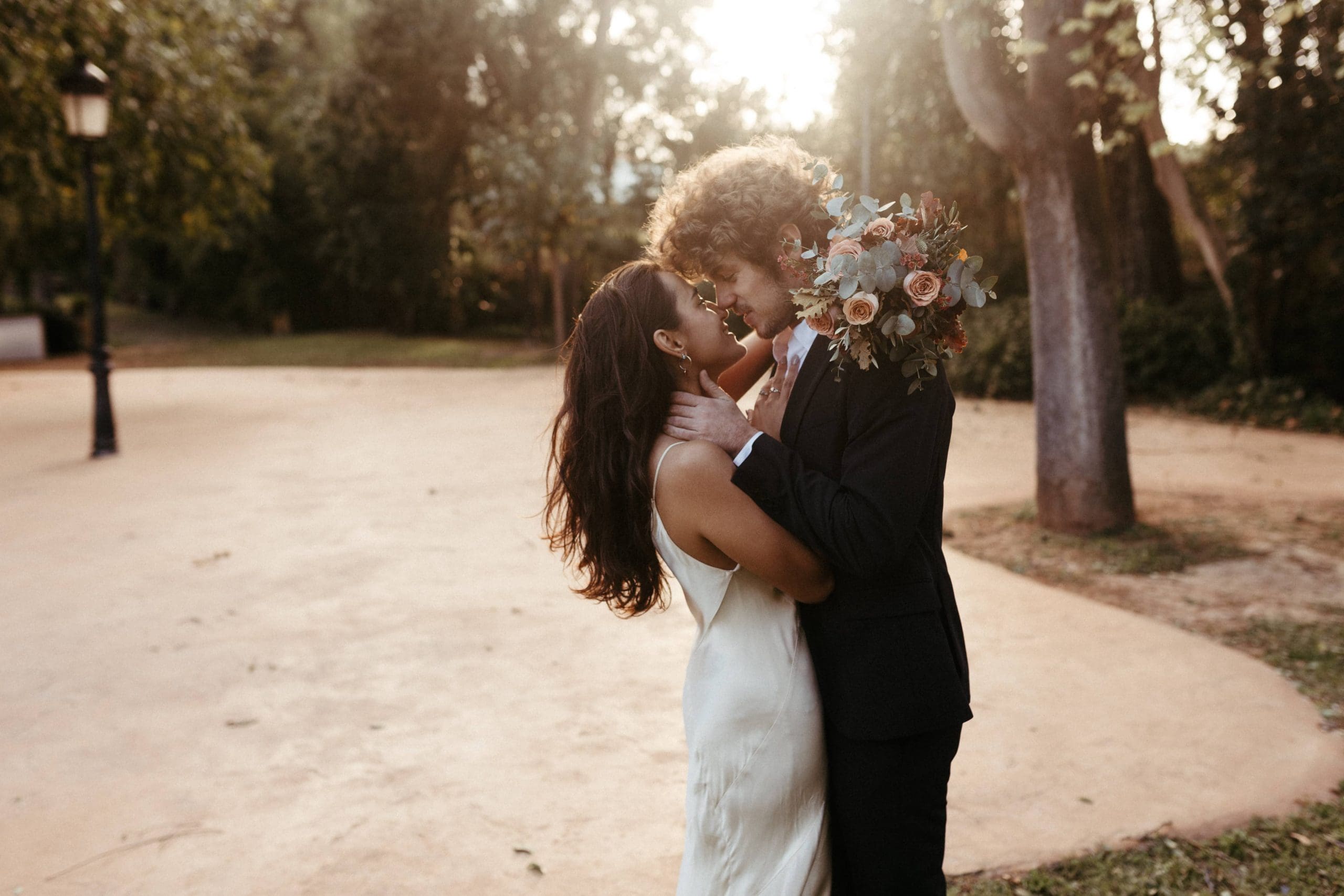 Pareja de novios besándose al atardecer en un paraje natural con árboles, con la novia sosteniendo su ramo de novia, creando una escena romántica.