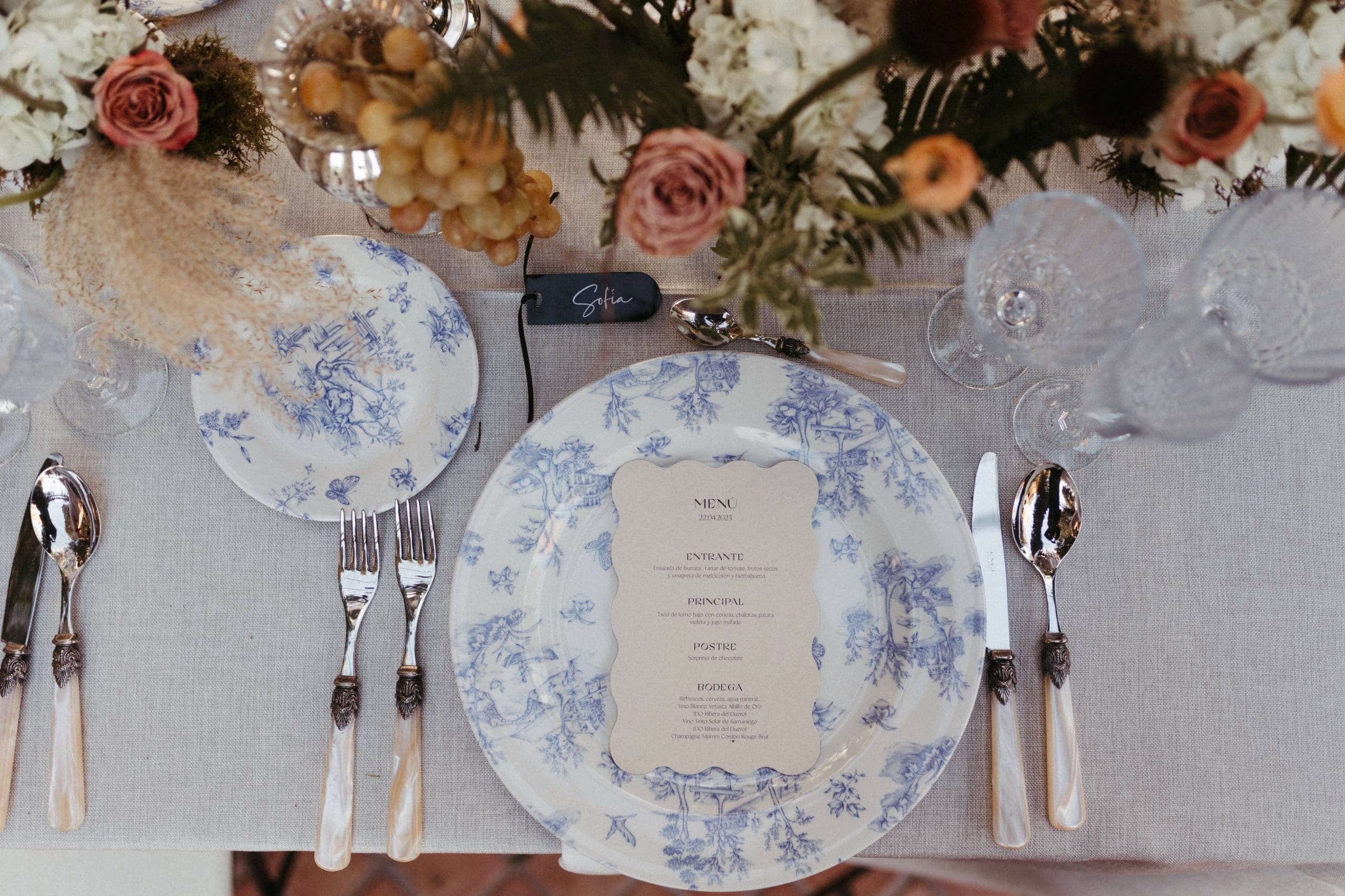 Mesa de boda decorada por Bouclé Weddings, con vajilla en tonos azules, cubertería de plata, motivos florales y uvas.