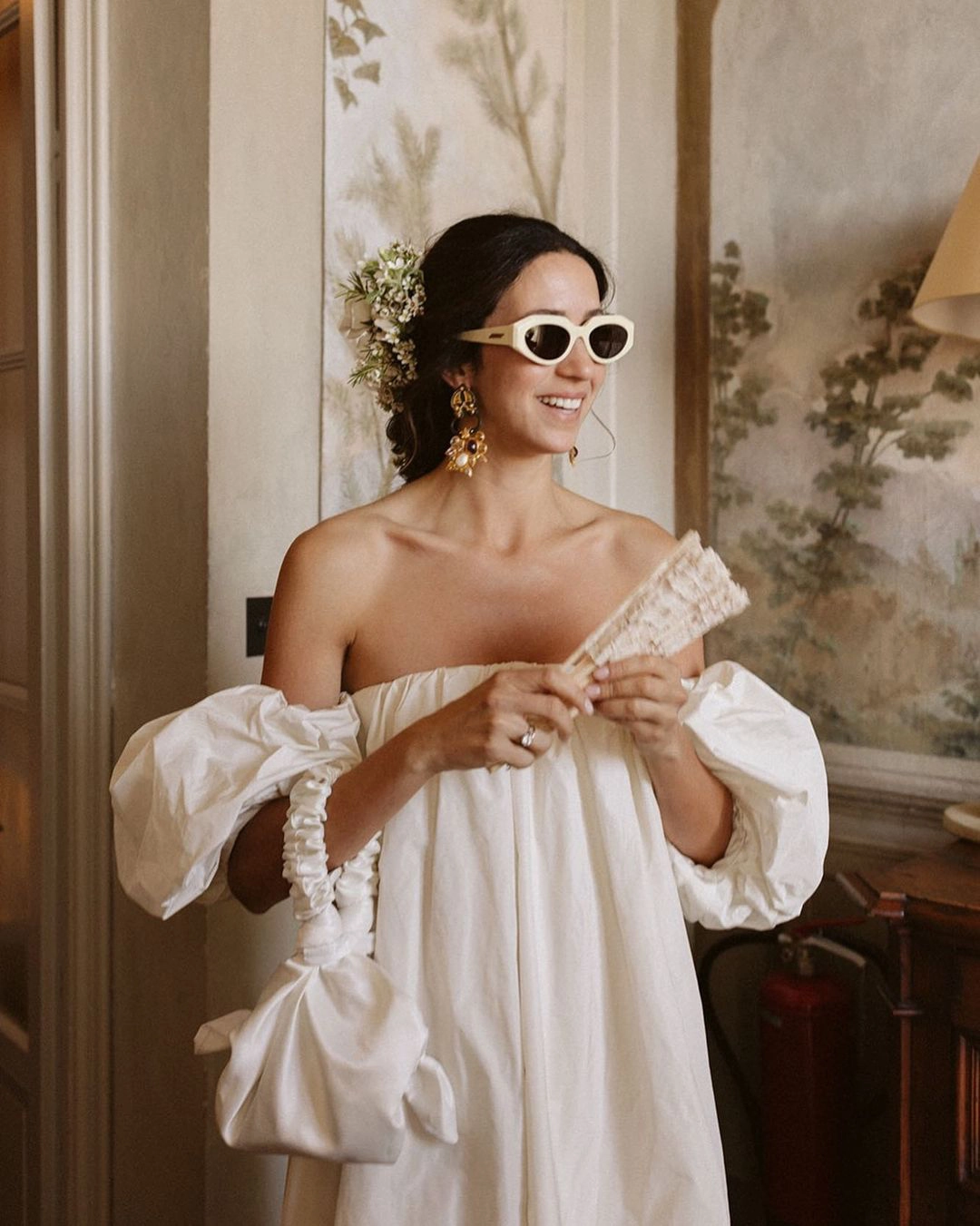 Novia con gafas de sol, abanico y bolso blanco a juego, deslumbrante en su boda.