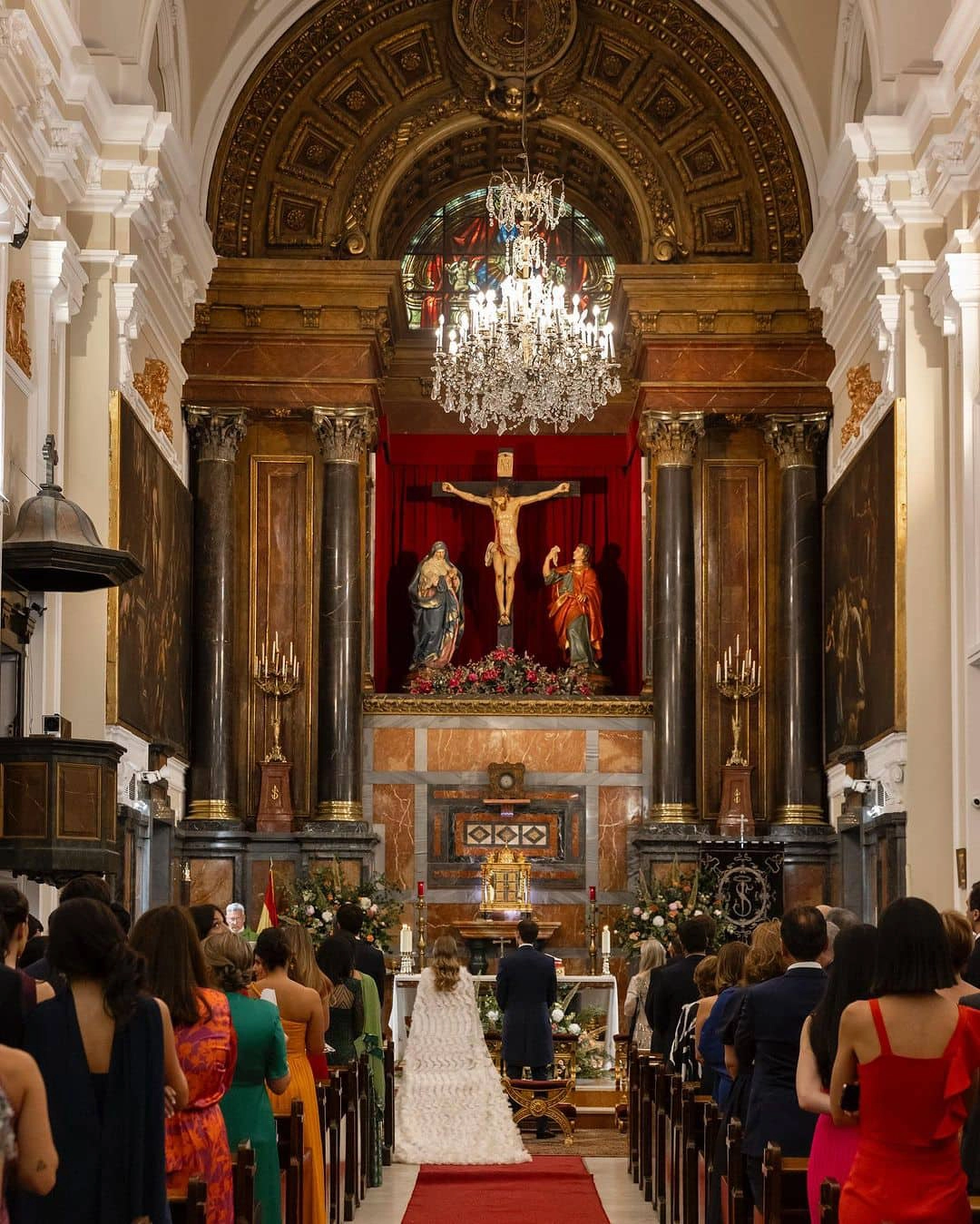 Vista completa de la iglesia desde la parte trasera, con invitados y novios en el altar mirando a la figura de Jesús durante el rito de la velación - Bouclé Weddings