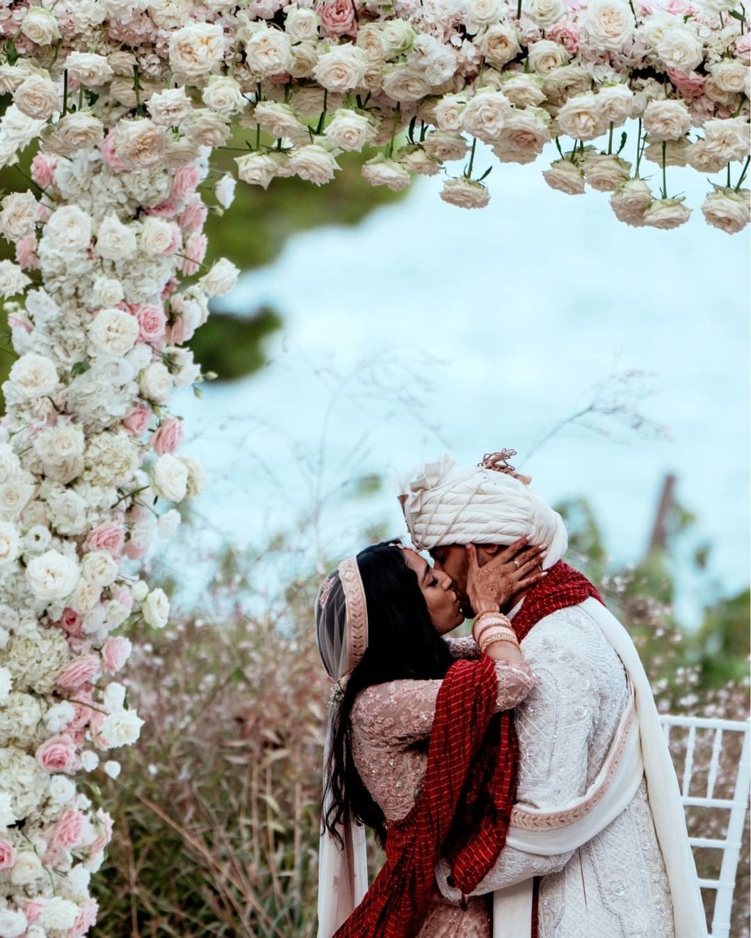 Pareja de novios hindúes compartiendo un beso romántico en Giardini del Fuenti, con un fondo pintoresco de la Costa Amalfitana, simbolizando la unión de culturas y amor.