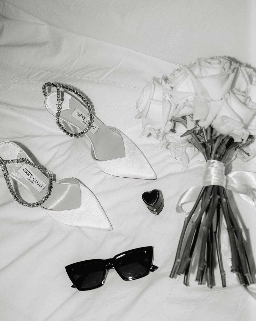 Zapatos de novia blancos, gafas de sol estilizadas y un frasco de perfume elegante dispuestos sobre una cama con sábanas blancas.