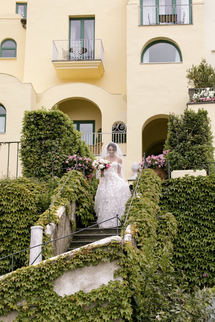 Novia descendiendo elegantemente las escaleras del Hotel Belmond Caruso, con el paisaje de la Costa Amalfitana de fondo, destacando la arquitectura histórica y el entorno romántico