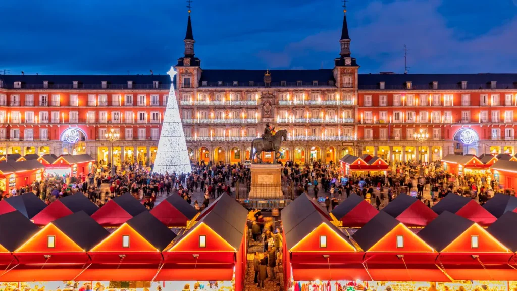 Mercadillo Navideño en Plaza Mayor, Madrid, con puestos de madera y decoraciones luminosas reflejando la tradición navideña española.