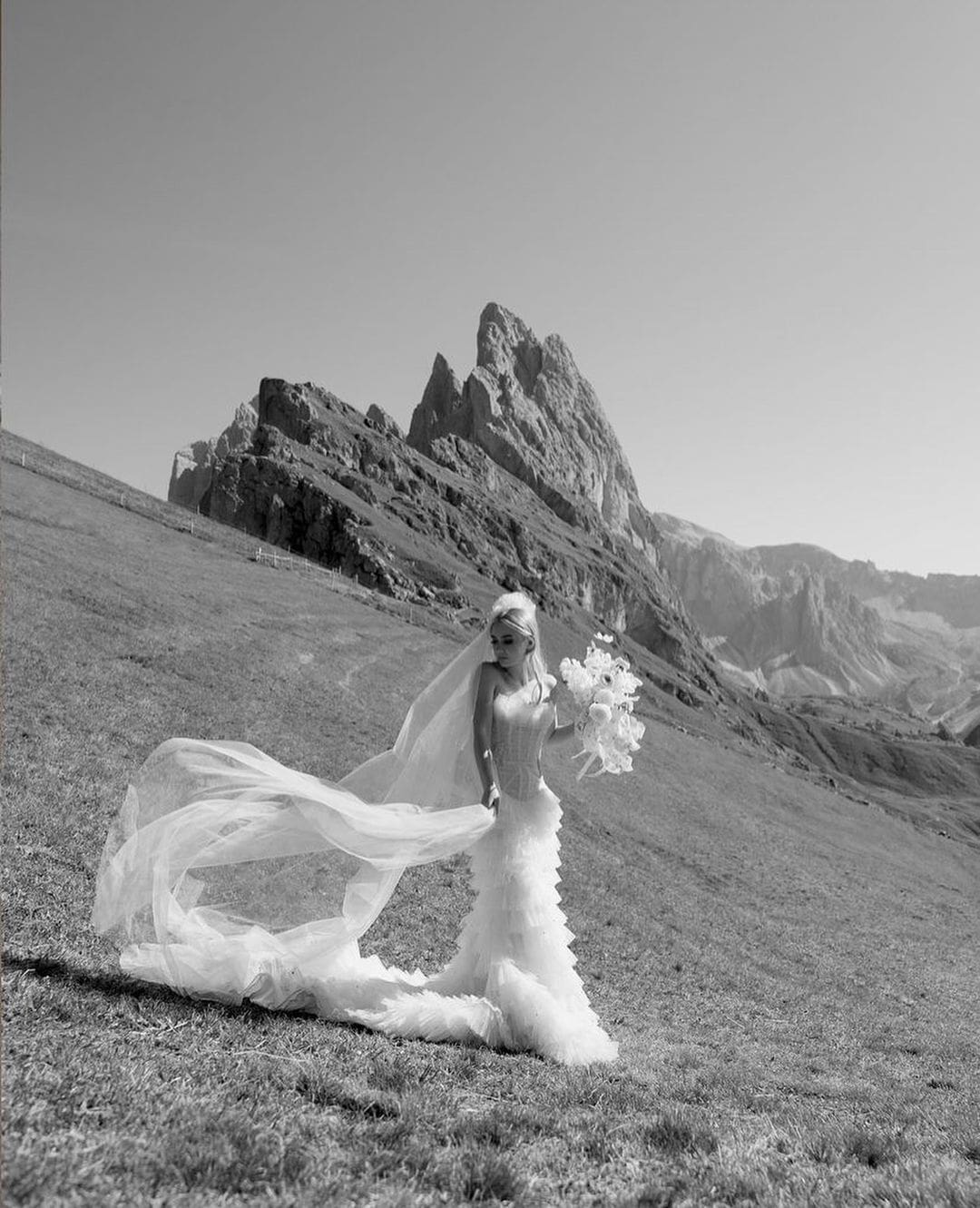 Novia posando en un campo de montaña, sosteniendo su ramo y su velo al viento.